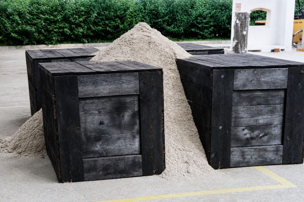 Vier schwarze Holzkisten kreuzförmig angeordnet, schräg von der Seite, in der Mitte ein Haufen Sand, der sich zwischen den Kisten verteilt