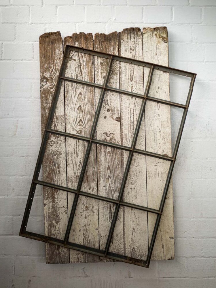 Altes rostiges Sprossenfenster-Gitter diagonal auf verwitterter, weiß gekalkter Holzbohlen-Wand montiert, an weißer Ziegelwand hängend