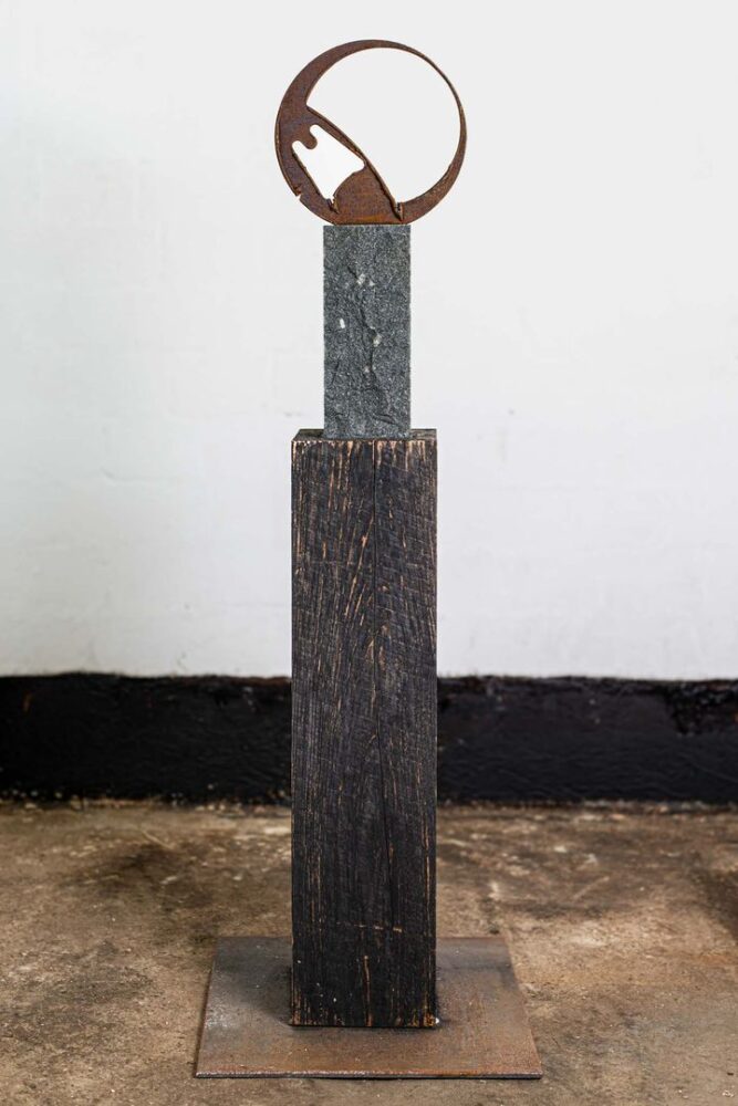 Skulptur mit rostigem Stahlring als Kopf, darunter eine schlanke Steinsäule auf einem dunklen Holzsockel mit Stahlplatte.