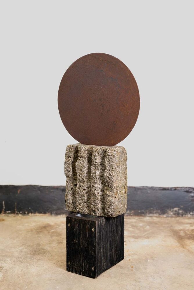 Skulptur aus rostiger Stahlscheibe auf grob behauenem Steinblock und dunklem Holzsockel, leicht seitliche Ansicht.
