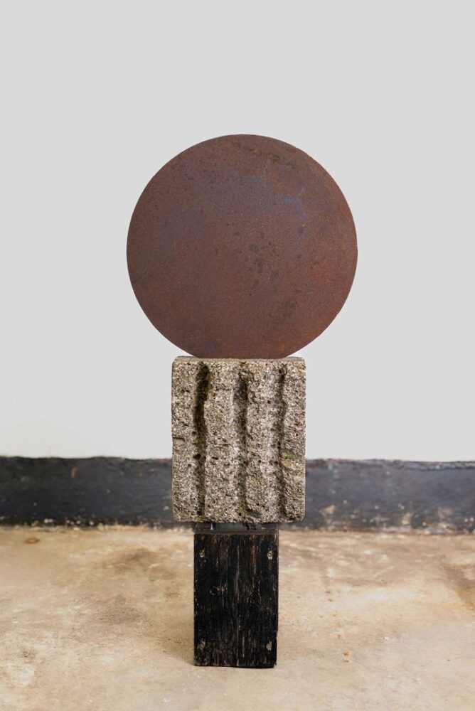 Skulptur aus rostiger Stahlscheibe auf grob behauenem Steinblock und dunklem Holzsockel, frontal betrachtet.