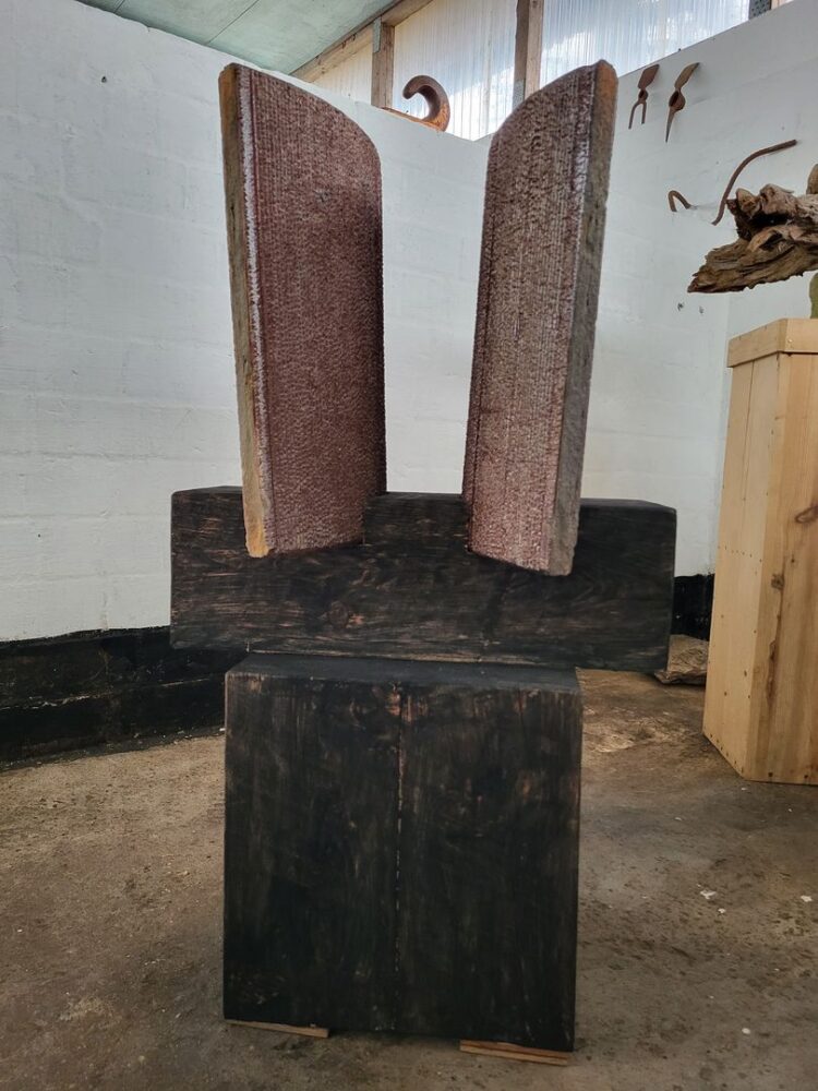 Skulptur mit zwei aufrecht stehenden, geschwungenen Elementen in rostrotem Ton auf gestapelten dunklen Holzblöcken, vor weißer Ziegelwand